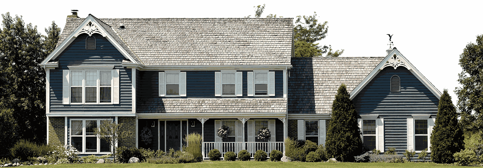 A large, two-story blue house with white trim and a grey shingle roof. It’s surrounded by shrubs, bushes, and trees, set against a black background.